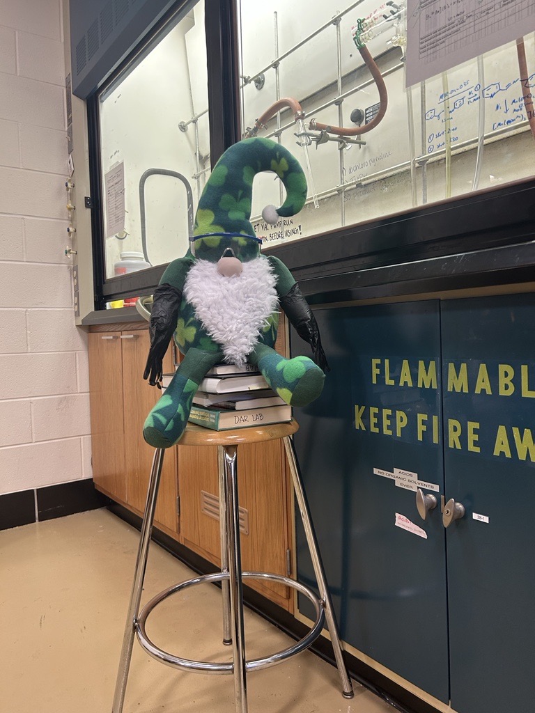 Jerome the leprechaun gnome sat in top of books placed in a stool wearing black latex gloves and goggles next to a chemistry workspace.