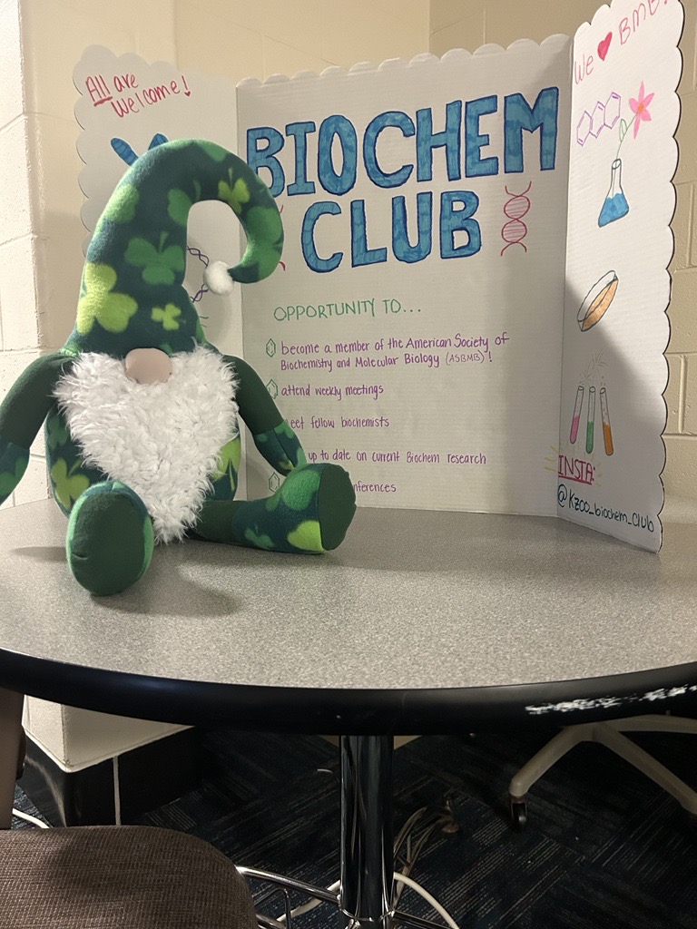 Jerome the leprechaun gnome sat on a table next to a trifold poster with information about the BioChem Club